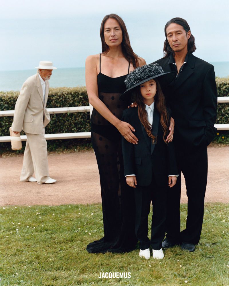 Family portrait of three models in black attire standing outdoors with the ocean and a white fence in the background.