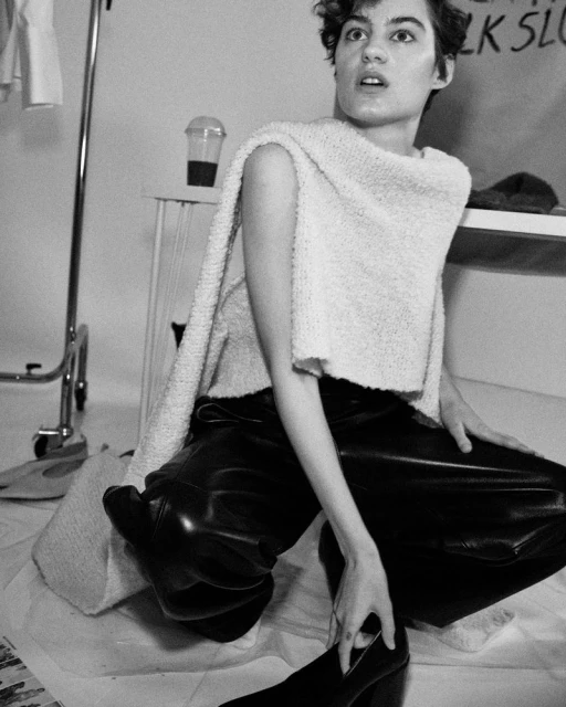 Monochrome shot of a young woman in a sweater and shiny pants, kneeling indoors. Coffee cup and clothing rack in the background.