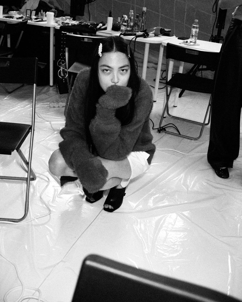 A person in an oversized sweater and shorts crouches indoors in a monochrome photograph. The setting appears to be a backstage area.