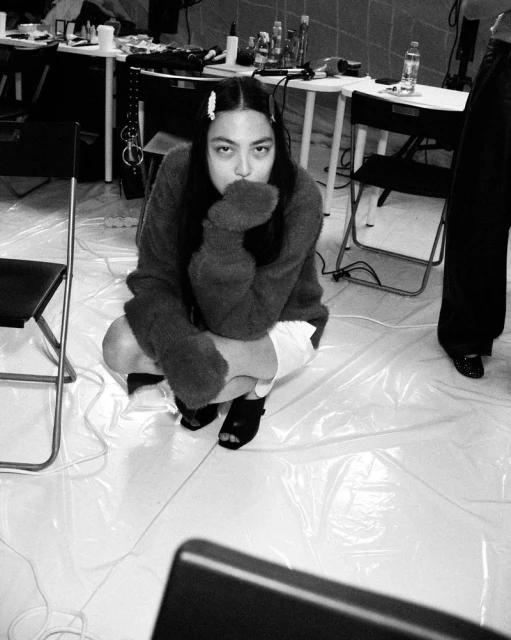A person in an oversized sweater and shorts crouches indoors in a monochrome photograph. The setting appears to be a backstage area.