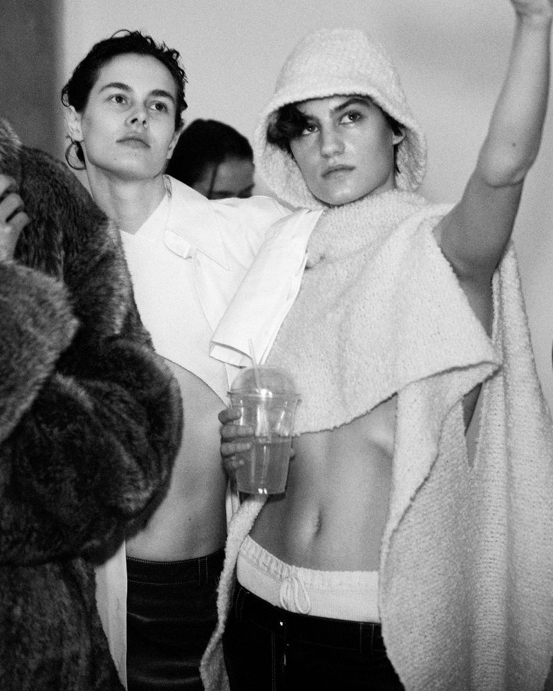 Black and white shot of women in fashionable clothing, one in a furry coat and the other in a hooded poncho, posing indoors.