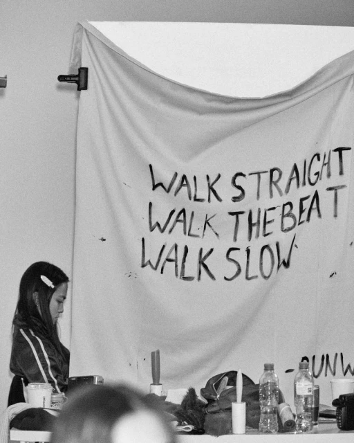 A young adult female stands near a table with water bottles, with a hand-painted sign hanging behind her in a monochromatic shot.