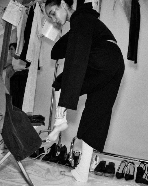 Black and white studio shot of a young adult female model in a black hooded sweat suit adjusting a white sock, with shoes and clothes rack.