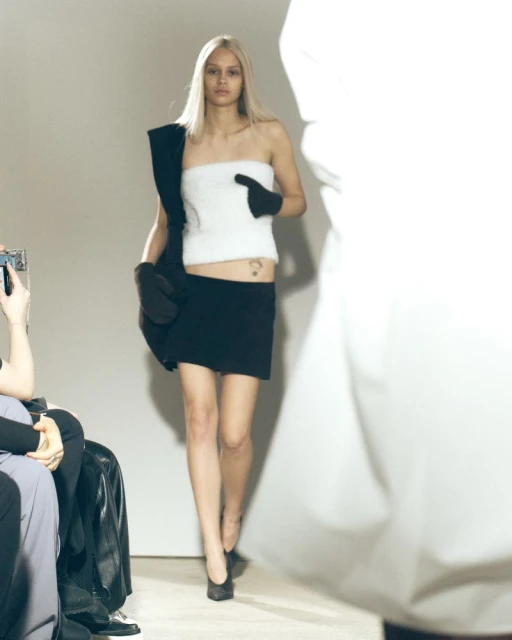 Fashion runway presentation featuring a blonde woman in a black skirt and white top walking past seated attendees in an indoor venue.