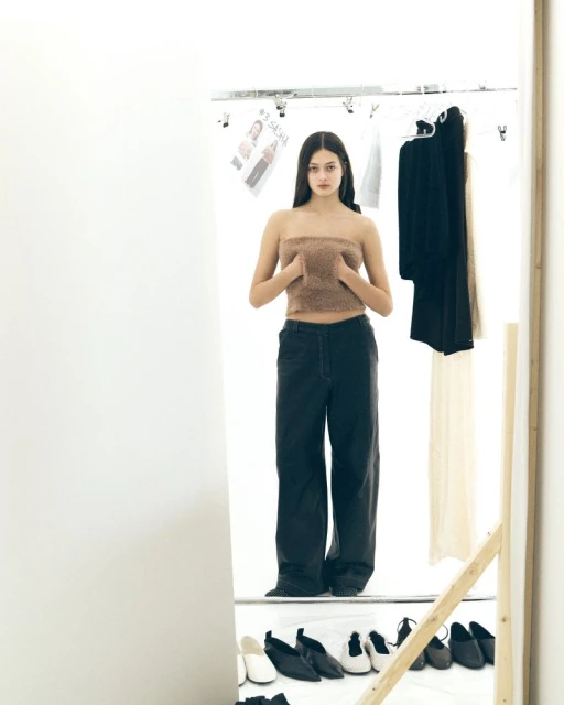 A young woman poses in a cropped top and dark pants, reflected in a mirror, in front of a clothing rack in a white studio.