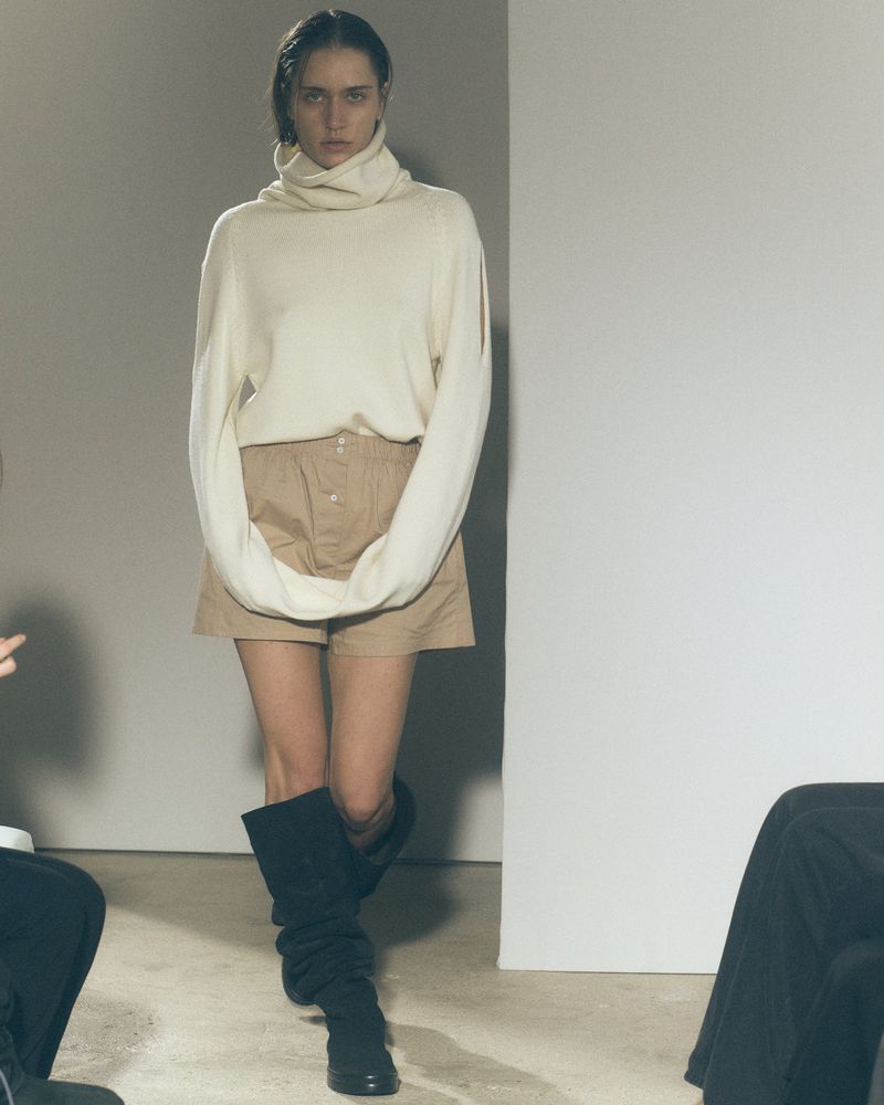 Fashion model walks runway wearing a white turtleneck sweater, khaki shorts, and black boots against a white backdrop.
