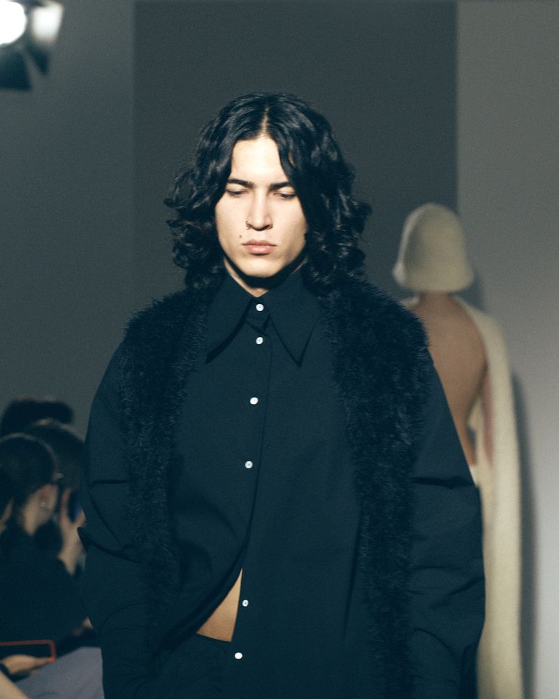 Runway fashion show featuring a model in a black collared shirt and furry black vest, walking with eyes closed and other models in background.