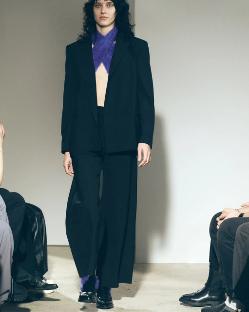 Runway model in black blazer and wide-leg pants walks during fashion show. Purple knit scarf and black patent leather shoes visible.
