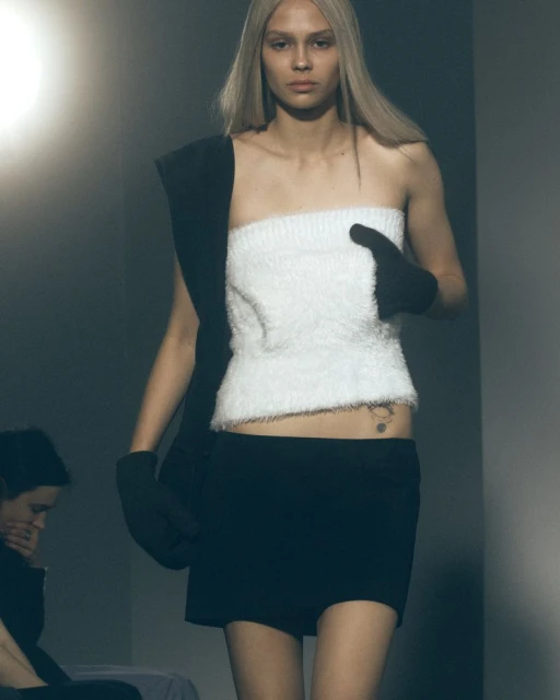 A blonde fashion model presents a striking pose in a white tube top and black mini skirt, accentuated by black gloves. Fashion show scene.