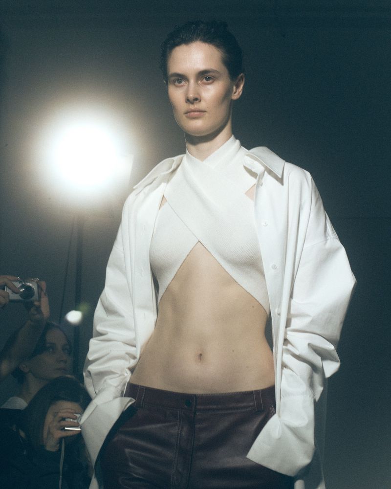 Fashion model on runway wearing white crop top and shirt with burgundy pants, posing in a studio with bright lighting.