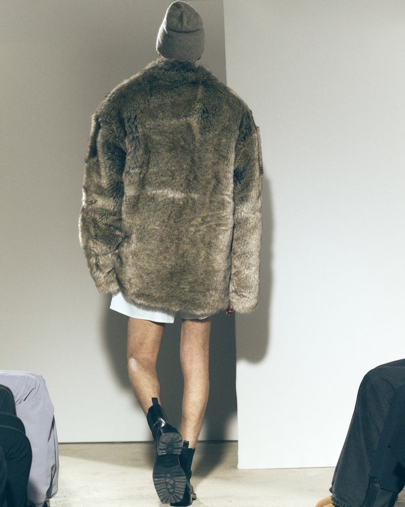 Back view of a person in a brown faux fur coat, beanie, white shirt, and black boots, standing in a brightly lit indoor space.