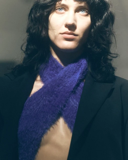 Close-up of a woman with blue eyes and dark, wavy hair wearing a purple scarf and black jacket, posing in a studio setting.