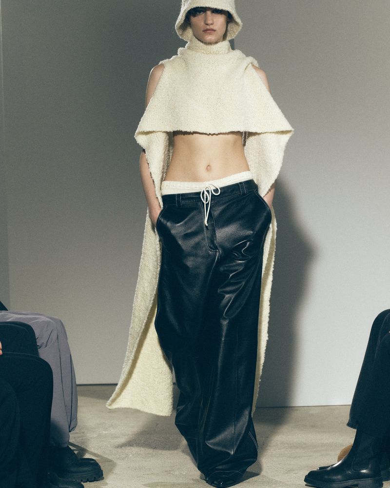 Fashion shot of a slender adult female model wearing a cream-colored knit hat, a top with cape sleeves, and baggy black faux-leather pants.