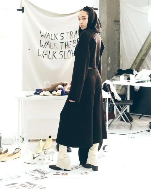 A woman in a dark brown hooded outfit poses indoors, with a white backdrop featuring painted black text, and shoes scattered on the floor.
