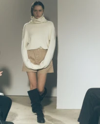 Model in cream turtleneck sweater and khaki shorts wearing tall black boots on a plain light background