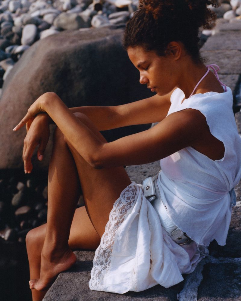Woman in a white dress sits in sunlight on rocky beach with stone boulders. She is barefoot and looking down in a contemplative pose.