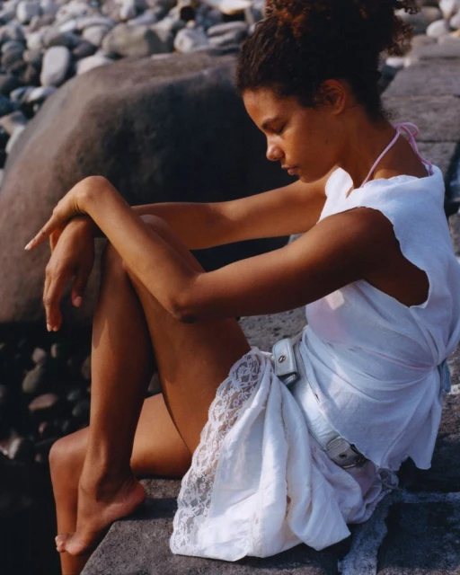 Woman in a white dress sits in sunlight on rocky beach with stone boulders. She is barefoot and looking down in a contemplative pose.