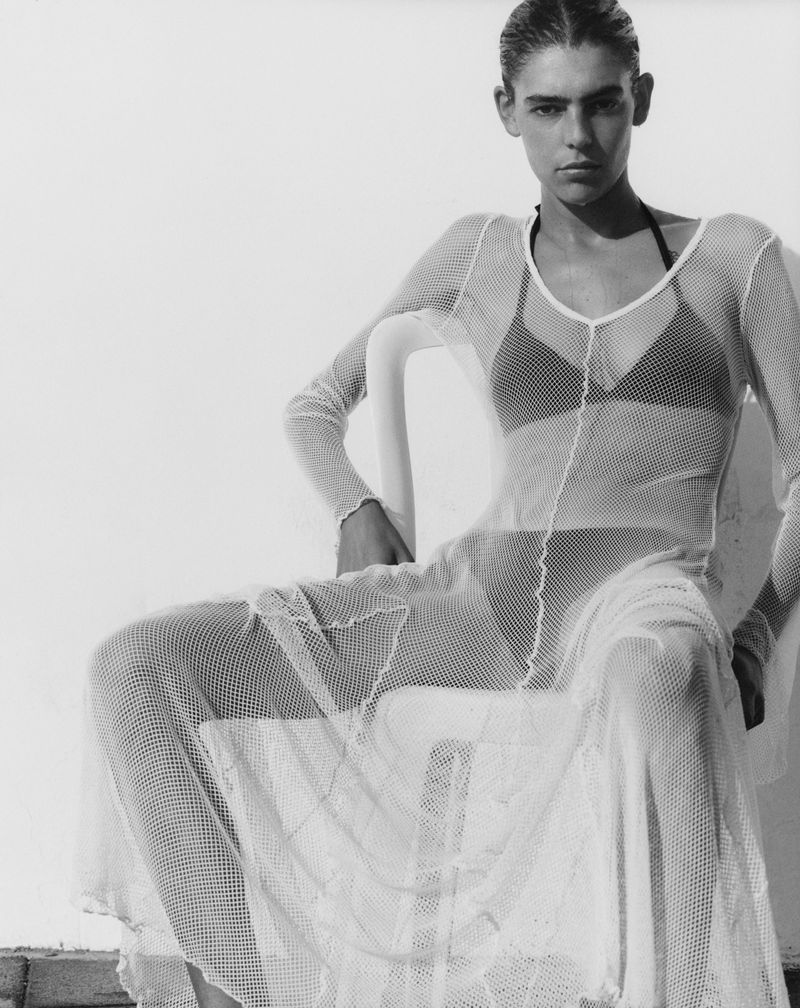 A lone female model in a full-length white mesh dress is seated outside against a light wall, her expression neutral and relaxed.