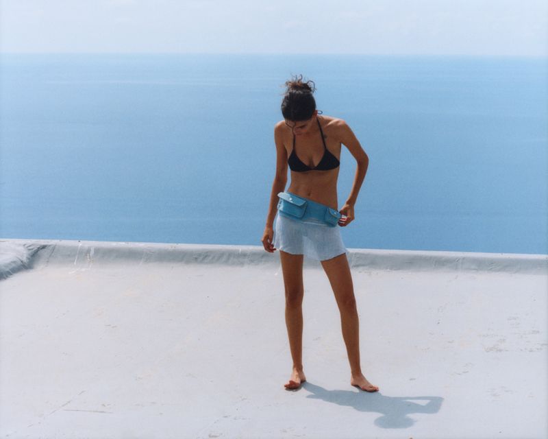 Adult female in a black bikini and light blue skirt stands on a rooftop overlooking the ocean on a bright sunny day.