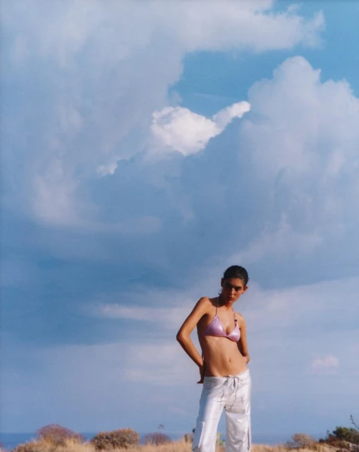 Young woman in a bikini top and white pants poses outdoors against a blue sky with clouds and dried plants at her feet.
