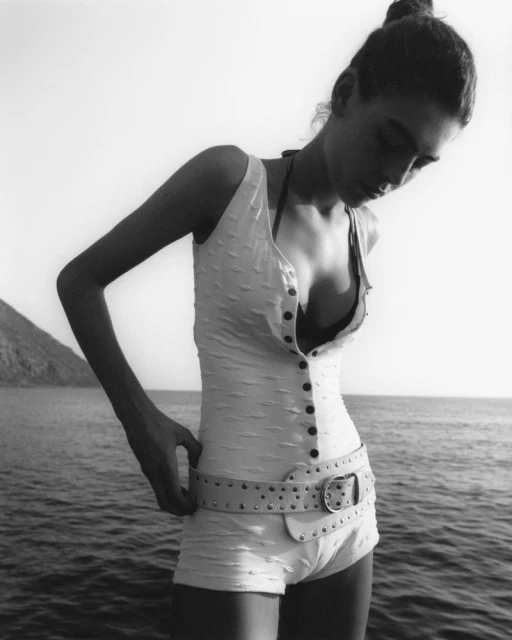 Monochrome image of a woman in a bathing suit with a wide belt, standing near the sea with a mountain in the background, looking down.