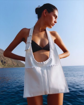 Fashion shot of a woman in a black bikini and white bag dress, standing near the sea on a sunny day. Blue sky and water background.
