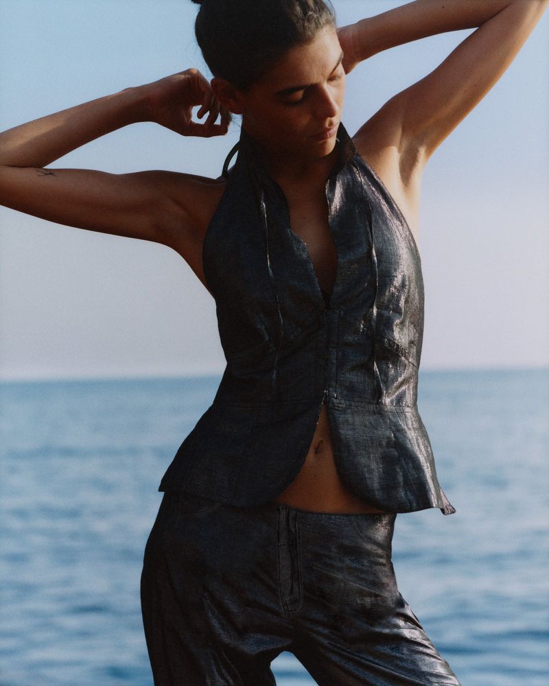 Woman in shimmering dark halter top and pants stands outdoors near the sea. Hands are behind her head, in a medium, portrait shot.