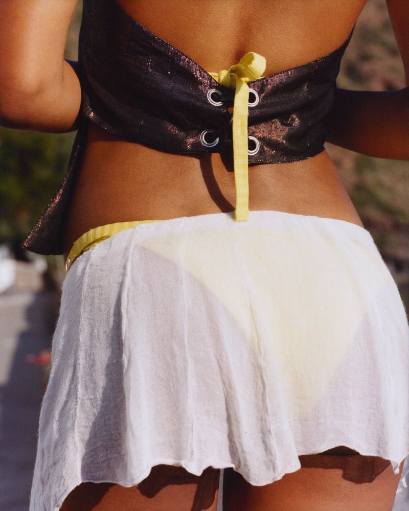 Back view of a woman wearing a brown halter top and a white skirt, posing outside with blurred background in a fashion shot.