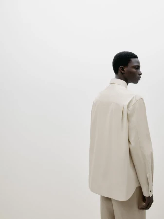 An African-American man in a light beige button-down shirt and pants is shown in a minimalist fashion shot against a white backdrop.