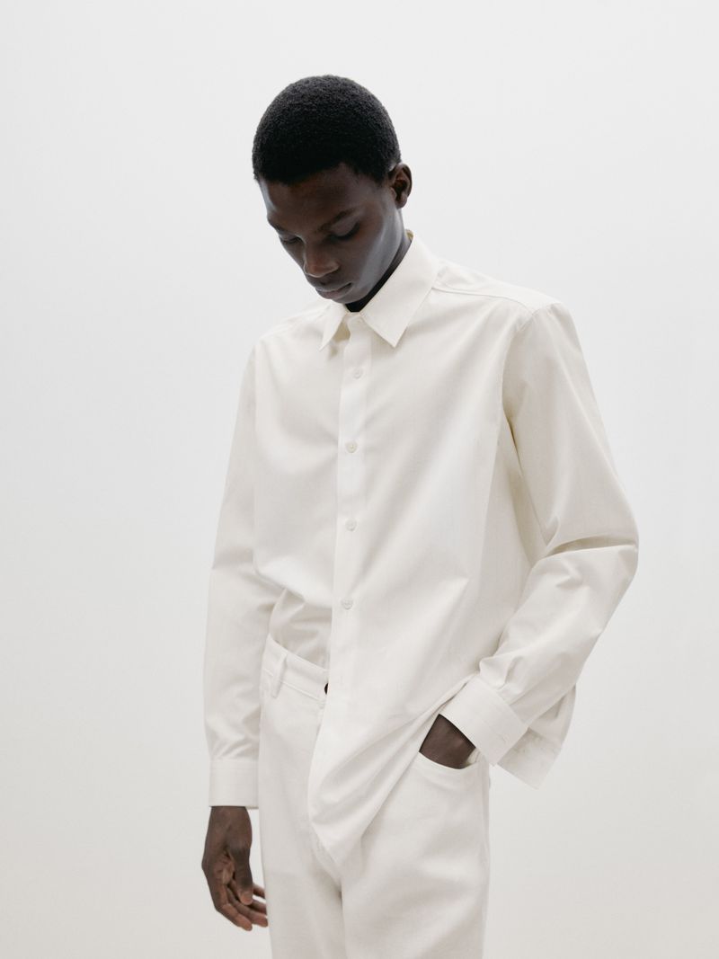 An African American man wearing an oversized, long-sleeved white shirt and matching pants stands indoors against a bright white background.