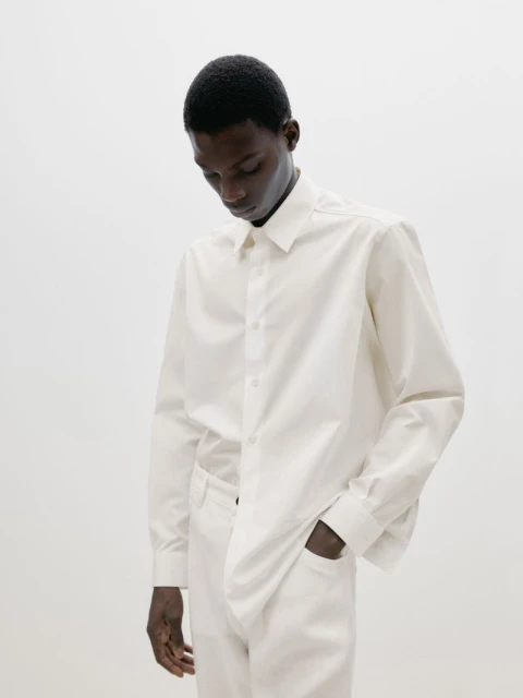 An African American man wearing an oversized, long-sleeved white shirt and matching pants stands indoors against a bright white background.