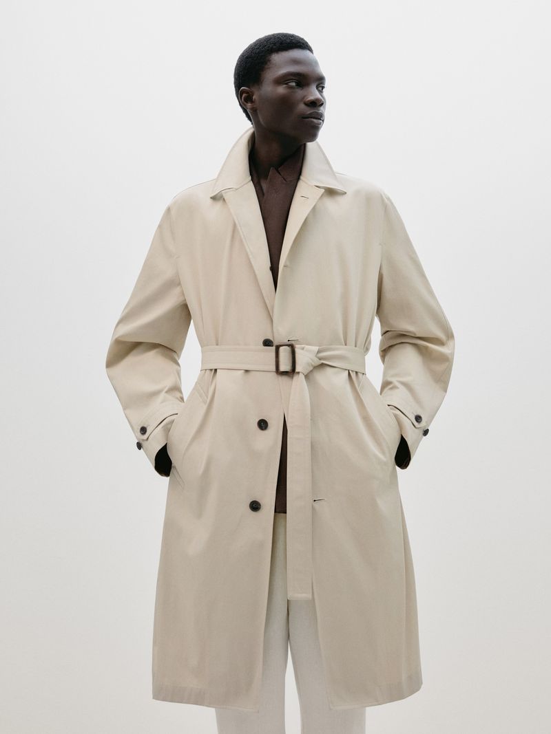 An adult male model poses in a beige trench coat and white pants against a neutral studio backdrop, showcasing classic menswear.