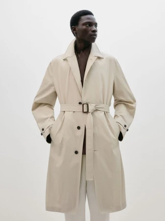 An adult male model poses in a beige trench coat and white pants against a neutral studio backdrop, showcasing classic menswear.