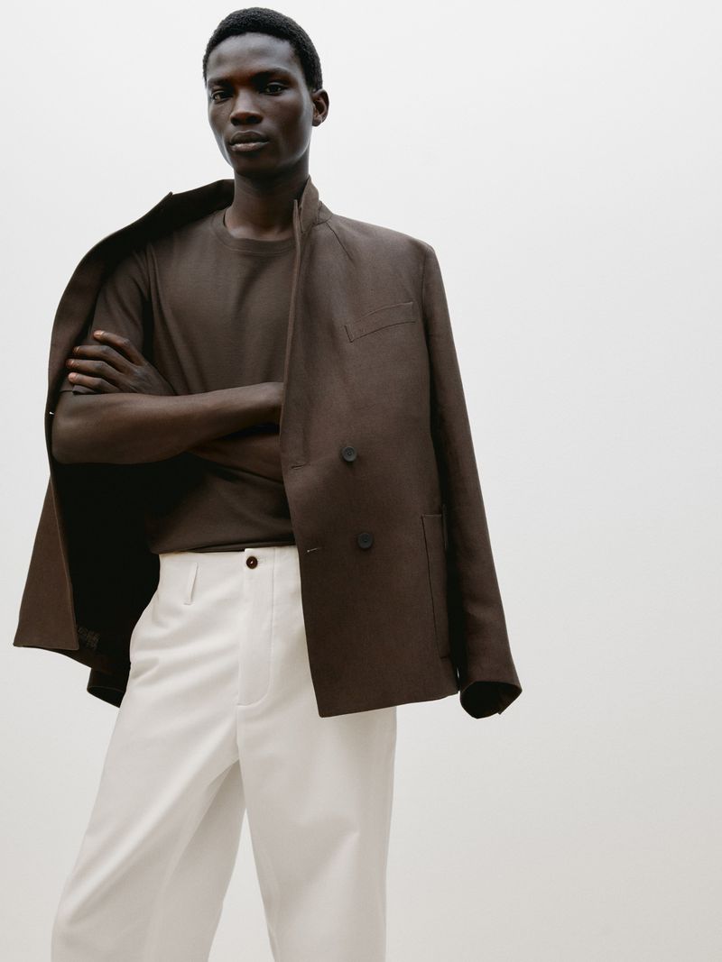 Dark-skinned man in brown t-shirt, white pants, and blazer jacket in a fashion studio portrait with arms crossed and a clean backdrop.