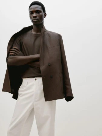 Dark-skinned man in brown t-shirt, white pants, and blazer jacket in a fashion studio portrait with arms crossed and a clean backdrop.