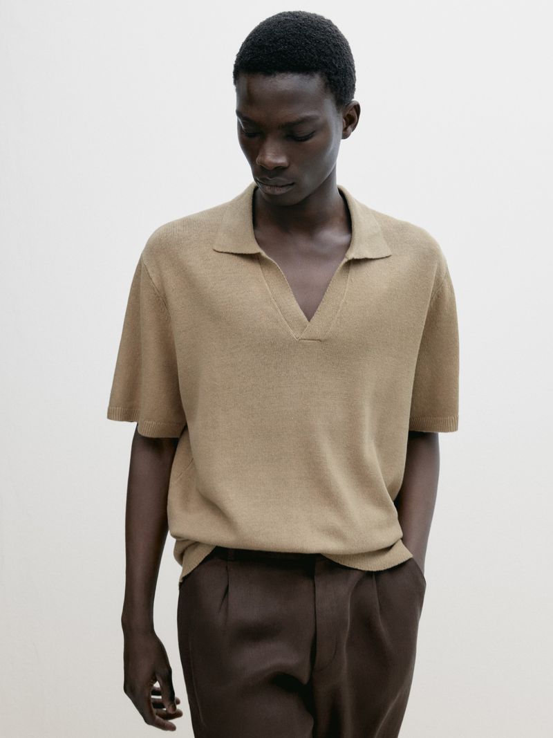 A single dark-skinned male is photographed indoors wearing a tan short-sleeved knit polo shirt and dark dress pants against a plain white background.