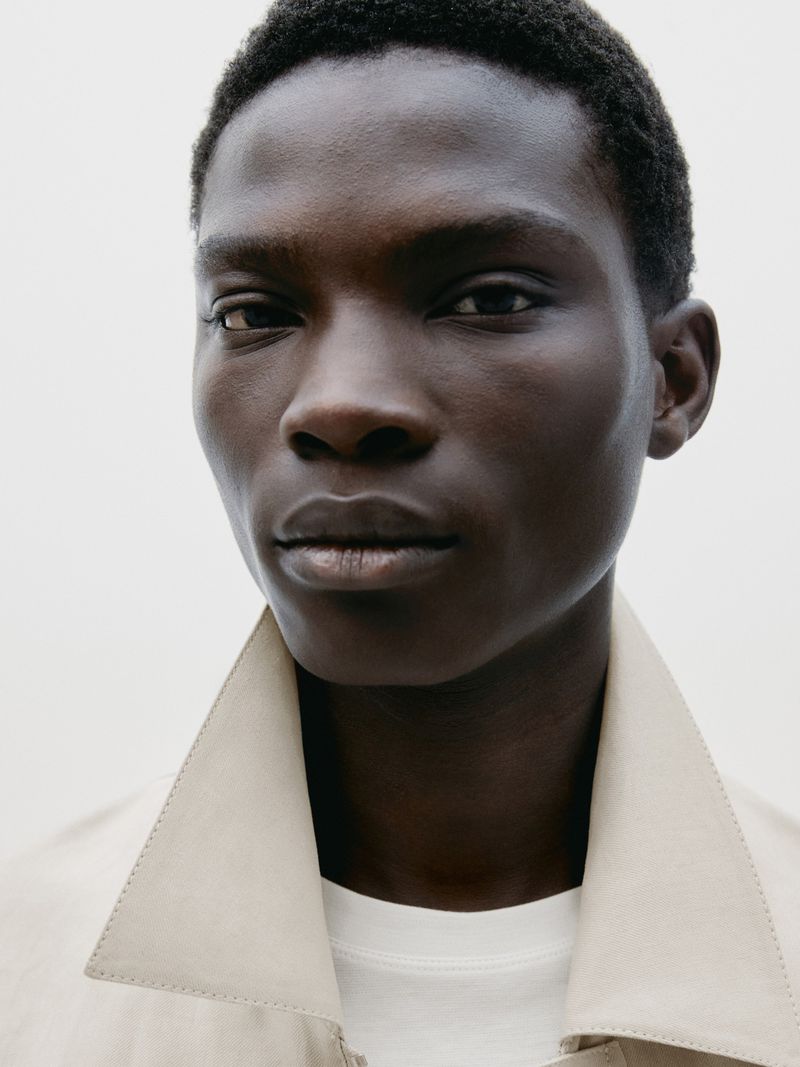 Close-up of a Black man with short hair wearing a beige trench coat against a white background, looking directly at the camera.