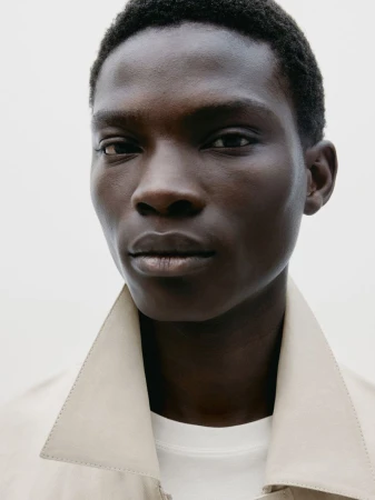 Close-up of a Black man with short hair wearing a beige trench coat against a white background, looking directly at the camera.