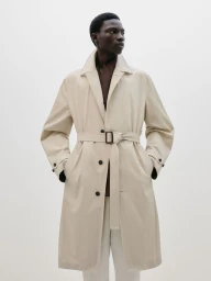 Model wearing a beige belted trench coat and white trousers against a plain white studio background.