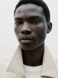 Close-up portrait of a black man wearing a light beige collared jacket against a plain white background