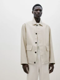 Black man wearing cream button-up collared jacket and matching trousers against a plain white studio background