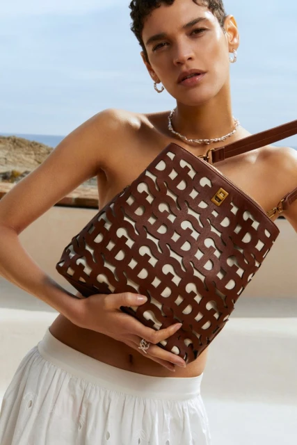A woman with short hair poses outdoors with a brown and white tote bag, wearing a white skirt, hoop earrings, and a beaded necklace.