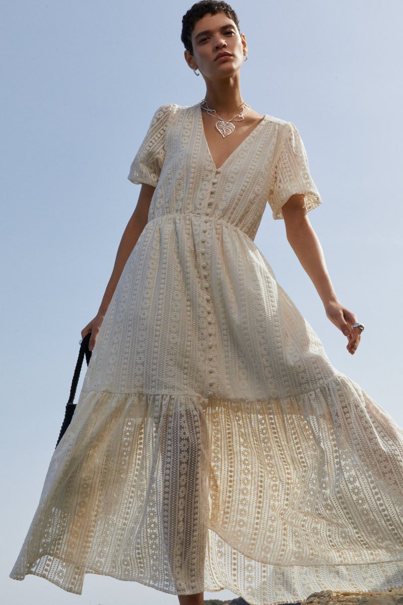 Woman in a patterned white dress and jewelry standing outside against a clear blue sky. Low angle, upward perspective.