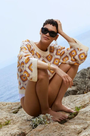 A woman with short hair and sunglasses poses on rocks near the ocean wearing a patterned sweater and jewelry in an outdoor fashion shot.