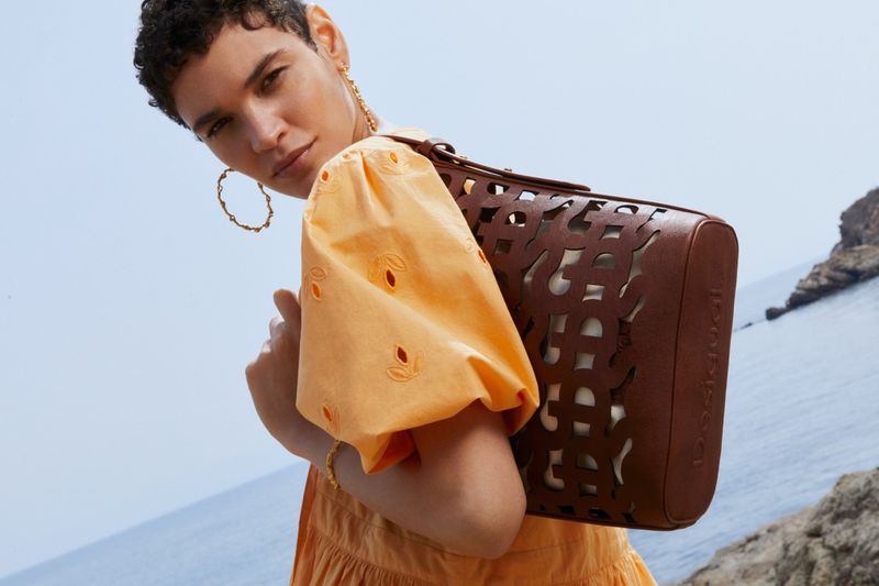 A light-skinned woman wearing an orange dress and gold hoop earrings holds a brown handbag in front of a coastal landscape.