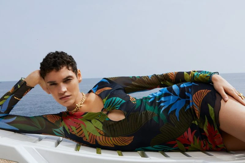 Adult woman in a tropical-print dress is lying on a white lounge chair outdoors under a blue sky next to the ocean.