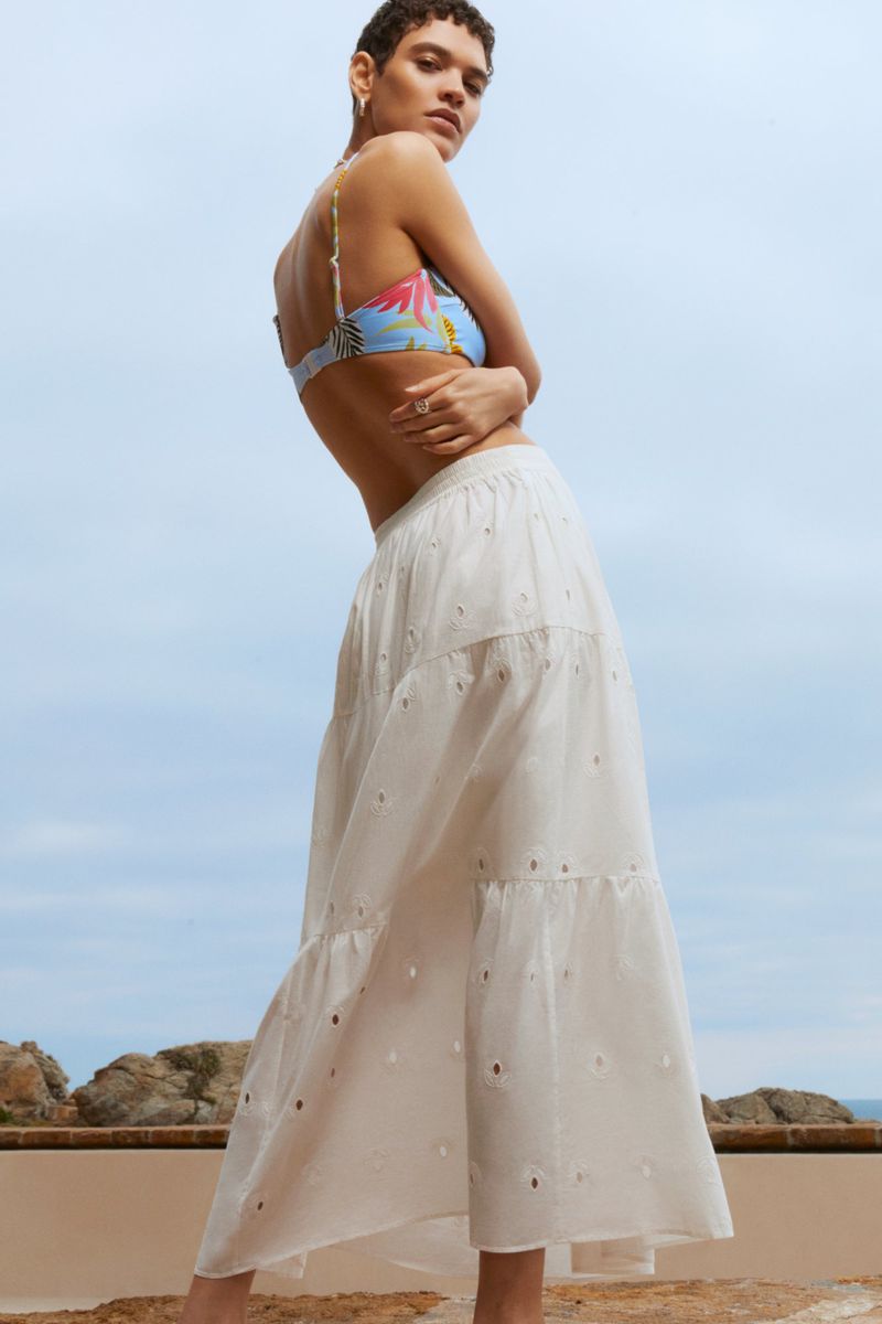 A woman models a floral bikini top and an eyelet skirt against a blue sky with rocky landscape in the background, shot at eye-level.