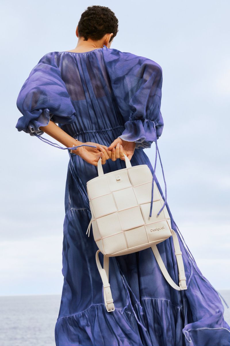 Adult woman from the back holding a white woven leather backpack, wearing a long blue dress outdoors against a light blue sky.