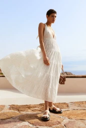 Model with short dark hair wearing a long white eyelet halter dress and sandals against a coastal landscape background.