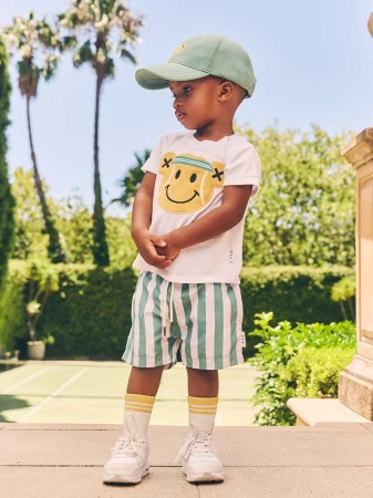 A young boy stands outside in a white t-shirt and striped shorts, accessorized with a green cap and white sneakers on a sunny day.